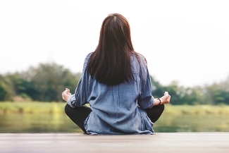 An image of a woman in the lotus yoga pose, facing away looking at a lake.